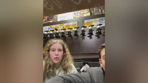 Media: Video of a blonde girl with curly hair, wearing a white sweater, sitting in a dimly lit restaurant, surrounded by toy animals hanging on a dark wall.