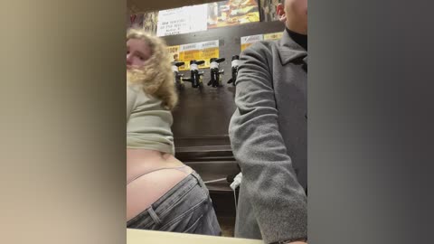 Media: Video of a woman with curly blonde hair, wearing a light green shirt, and grey pants, bending over to grab a drink from a soda machine.