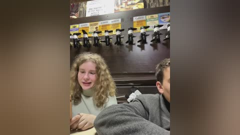 Media: A video captures a young girl with curly blonde hair and a light sweater chatting with a man in a gray sweater. They sit at a counter with a row of beer taps behind them.