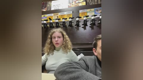 Media: A video shows a young woman with curly hair in a white sweater sitting at a bar, flanked by two men in gray and white shirts, with a background of beer taps and signs.