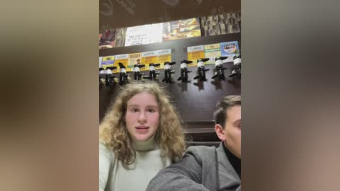 Media: Video of a young woman with curly blonde hair, wearing a light sweater, sitting in a dimly lit arcade. Behind her, black and white plastic penguin figures hang from a wall with colorful signs above.