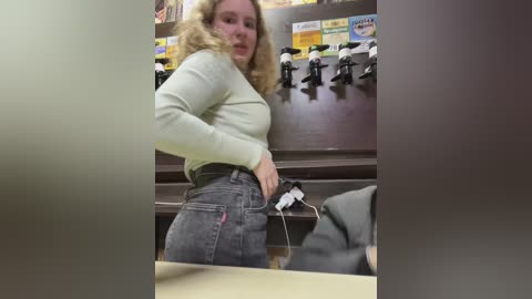 Media: A video shows a blonde woman with curly hair, wearing a light green sweater and high-waisted jeans, pulling down her pants in a store.