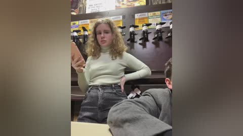 Media: Video of a blonde woman in a light green turtleneck, high-waisted jeans, holding a phone, standing in a toy store with shelves of action figures in the background.