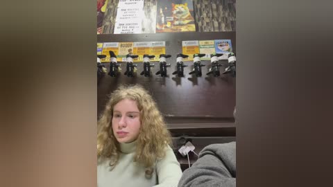 Media: Video of a young, curly-haired girl in a beige turtleneck sweater, sitting in a booth with a dark wooden background featuring toy figurines and a \"Bart Simpson\" sign.