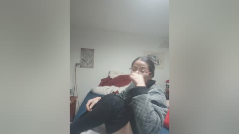 A video of a young woman with glasses, wearing a gray sweater and black pants, sitting on a bed with red and white bedding, in a dimly lit bedroom with white walls and abstract art.
