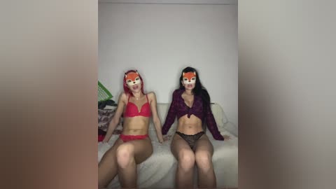 Video of two women in matching red fox masks and lingerie, sitting on a white couch. The background is plain white, with a laundry basket and clothes on the left.