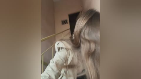 Media: A video captures a blonde woman with long, wavy hair standing on a staircase, partially obscured by the railing. The background features beige walls and a closed door.