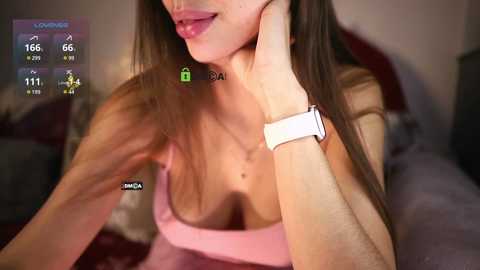 Media: Video of a fair-skinned woman with long brown hair, wearing a pink bra, resting her chin on her hand. Background shows a dimly lit room with a gray couch.