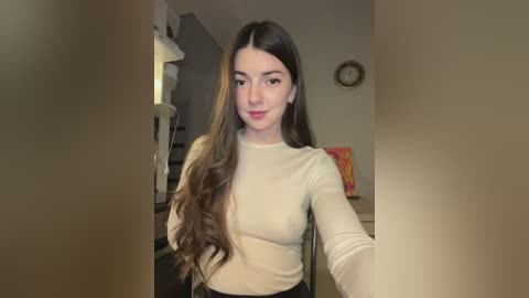 Media: Video of a young woman with long, wavy brown hair, fair skin, and a slender figure, wearing a fitted cream top. She's seated indoors, with a beige wall, a clock, and a shelf in the background.