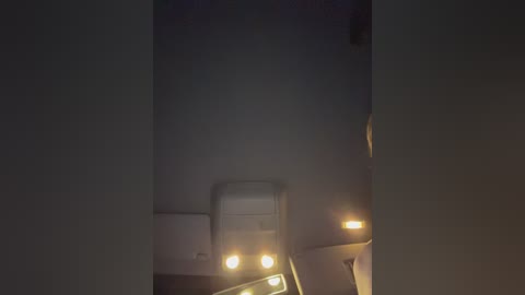 Media: Video of a dark, foggy night showing a white car with three bright headlights in the foreground, partially obscured by the dense fog. The background is dark and indistinct, creating a moody, mysterious atmosphere.