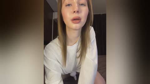 Media: Video of a young Caucasian woman with fair skin, straight light brown hair, and light blue eyes, wearing a white long-sleeve shirt, leaning forward slightly, in a dimly lit bedroom with dark curtains.