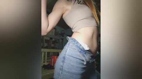 A video of a slender, light-skinned woman with long, straight, blonde hair, wearing a beige crop top and high-waisted, light blue jeans, standing in a dimly lit, cluttered room with a wooden crib and red bucket in the background.