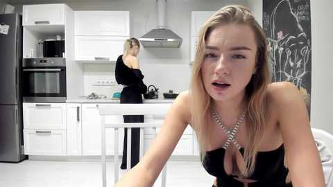 Media: Video of a blonde woman in a black strapless dress, sitting in a modern white kitchen, looking distressed. Another woman in a black top and skirt is cooking in the background.