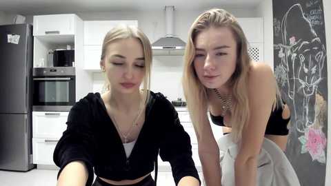 Media: Video of two blonde women in a modern kitchen. One wears a black jacket, the other a black sports bra, leaning on a counter. Background includes white cabinets, a black fridge, and a chalkboard wall with drawings.