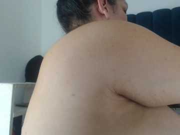 Video of a nude, overweight person with light skin, showing their back and upper arm, viewed from the side. They have short dark hair and are seated on a dark blue cushioned chair in a dimly lit room.