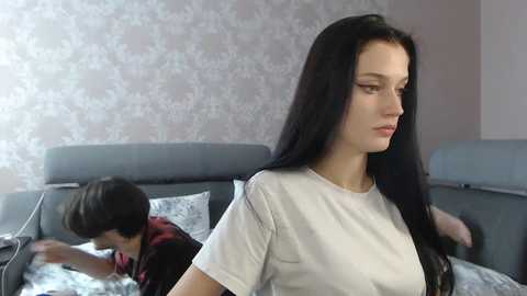 Media: Video of a young woman with long black hair and fair skin, wearing a white T-shirt, looking contemplative beside a gray couch with a child asleep on it. The background features a floral-patterned wallpaper.