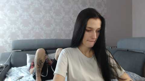 Media: Video of a young woman with long black hair, pale skin, and a small tattoo on her arm, sitting on a gray bed, in a room with floral wallpaper and a blue sofa.