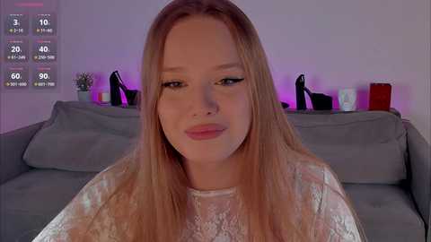 Video of a young, fair-skinned woman with long blonde hair, wearing a white lace top, seated on a grey sofa in a dimly lit room. Background includes a purple-lit shelf with a potted plant and high heels.