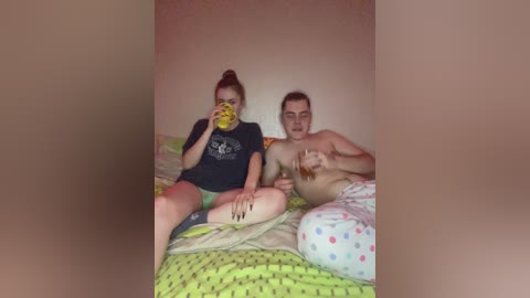 Video of a couple in bed, woman with a bun, drinking from a yellow cup, man shirtless, holding a brown bottle, colorful bedspread, beige wall, casual setting.