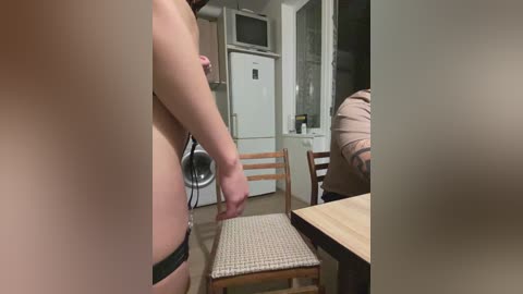A video shows a nude person with fair skin and a slender build, wearing black underwear, standing in a small kitchen. The background includes a white fridge, a microwave, and a wooden chair with a beige cushion.