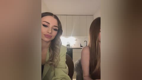 Media: Video of a slender, fair-skinned woman with wavy, shoulder-length brown hair, wearing a light green blazer, leaning forward in a modern kitchen with white cabinets and stainless steel appliances.