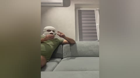A video of a man with a white mask and green shirt reclining on a light gray couch in a minimalist living room with white walls and a window with closed blinds.