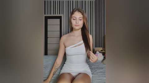 Video of a young, light-skinned woman with long brown hair, wearing a white, one-shoulder dress, sitting on a bed in a modern bedroom with gray and white striped wallpaper and a white dresser.