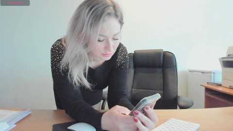 Video of a blonde woman in a black sweater, typing on a phone, seated at a wooden desk in a minimalist office.