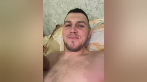 Video of a young, bearded Caucasian man with short brown hair lying shirtless on a bed with yellow and white striped pillows against a textured gray wall.