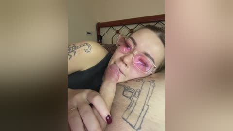 A close-up video of a light-skinned woman with pink glasses and black hair, lying on her side, licking a man's erect penis with a tattoo of a snake on her arm.