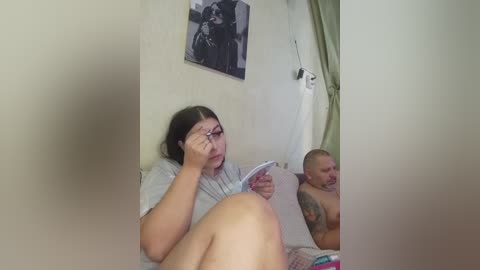 Video of a woman with glasses reading a magazine in bed, a man with a tattoo on his arm lying beside her, both shirtless, in a dimly lit room with a black and white framed photo on the wall.