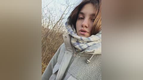 Media: A video of a young woman with long brown hair, wearing a light grey puffer jacket and a striped scarf, standing in a barren, outdoor winter landscape.