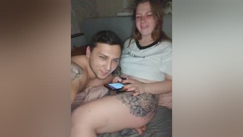 Video of a shirtless man with tattoos and a woman with long hair and a cropped top, playing on a smartphone in bed, blurred background.