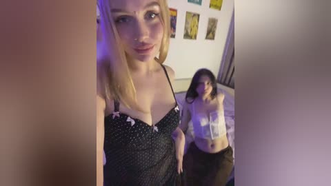 A video shows a blonde woman in a black polka-dot bra with pink bows, standing beside a topless woman with dark hair and a white crop top, in a dimly lit room adorned with colorful art.