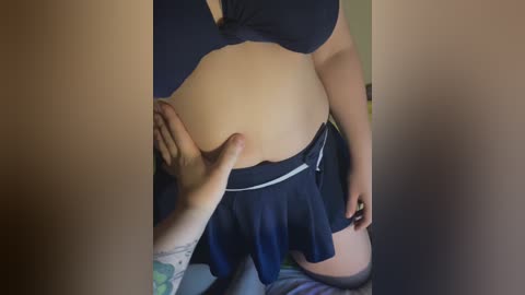 Video of a pregnant woman in a black bra and skirt, with a person's hand touching her stomach. Visible tattoos on the person's arm. Background shows a blurred indoor setting.