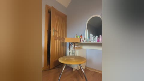 Media: Video of a simple, cluttered bathroom with a wooden door, a round mirror, a vanity counter with toiletries, and a small wooden stool.