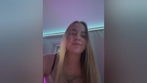 Media: A video of a young blonde woman in a beige lace bra, slightly out of focus, with a soft purple light in the background, creating a sensual, intimate atmosphere.