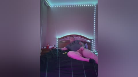 Media: A video of a plus-sized woman with fair skin, lying on a bed in a dimly lit bedroom with neon lights, wearing a grey top and no bottoms.