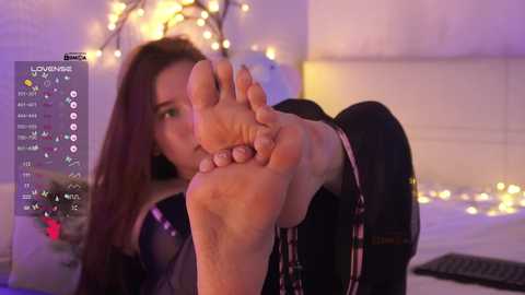 Video of a young woman with long brown hair, holding her bare foot in front of the camera, in a dimly lit bedroom with white sheets and fairy lights.