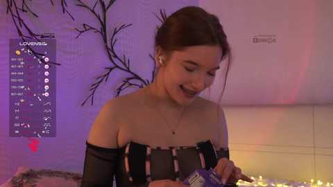 Video of a young woman with light skin and brown hair, wearing a black off-the-shoulder dress, smiling while pouring a drink. Background features a dimly lit, modern room with a purple wall and a Dymo label printer.