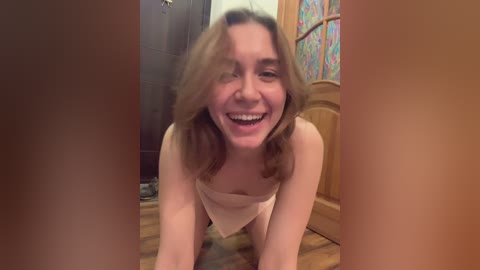 Media: Video of a smiling young woman with light skin and shoulder-length brown hair, wearing a towel, kneeling on a wooden floor in a bathroom with stained glass window and wooden door.