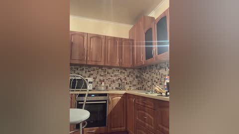 Media: Video of a modern kitchen with brown wooden cabinets, beige tiled backsplash, stainless steel appliances, a white stool, and a small round table.