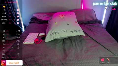 A video of a bed with a pillow that spells \"Lover\" in pink letters, surrounded by a laptop, a mouse, and a notepad, in a dimly lit room.