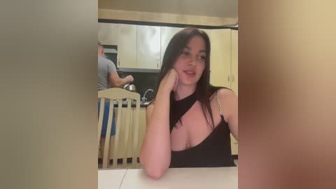 Media: Video of a young woman with long dark hair and a revealing black top, seated at a kitchen table, looking at the camera. A man is visible in the background, preparing food.