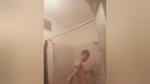 Video of a naked man with a tattooed arm, showering in a beige-tiled bathroom with a metallic shower curtain, under dim lighting.