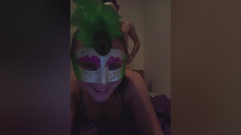 A dimly lit video shows a woman with a green feathered headpiece, purple eye makeup, and a white masquerade mask with a black jewel, smiling. She lies on a bed with dark sheets in a room with beige walls.