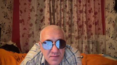 Media: Video of an older man with short white hair, wearing reflective sunglasses, resting his head on his hands in a bedroom with floral-patterned curtains.