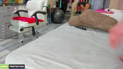 Media: A video of a messy bedroom with a white recliner chair, red pillow, brown pillow, and a TV remote on a gray bedspread. The background shows a cluttered room with a dresser, clothes, and a large exercise ball.