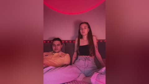 Media: Video of a young couple in a dimly lit room with red lighting, the man in a yellow shirt, the woman in a black crop top and high-waisted pants, both sitting on a bed.