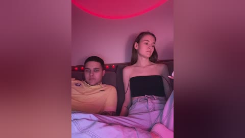 Media: Video of a young man and woman, both in casual attire, sitting on a bed under dim red lighting, appearing tense and distant.